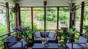 Ein überdachter Außenbereich mit einer Couch, zwei Sesseln und einem Couchtisch. Mehrere Kübelpflanzen umgeben die Sitzgruppe, und im Hintergrund ist ein Holzzaun zu sehen.