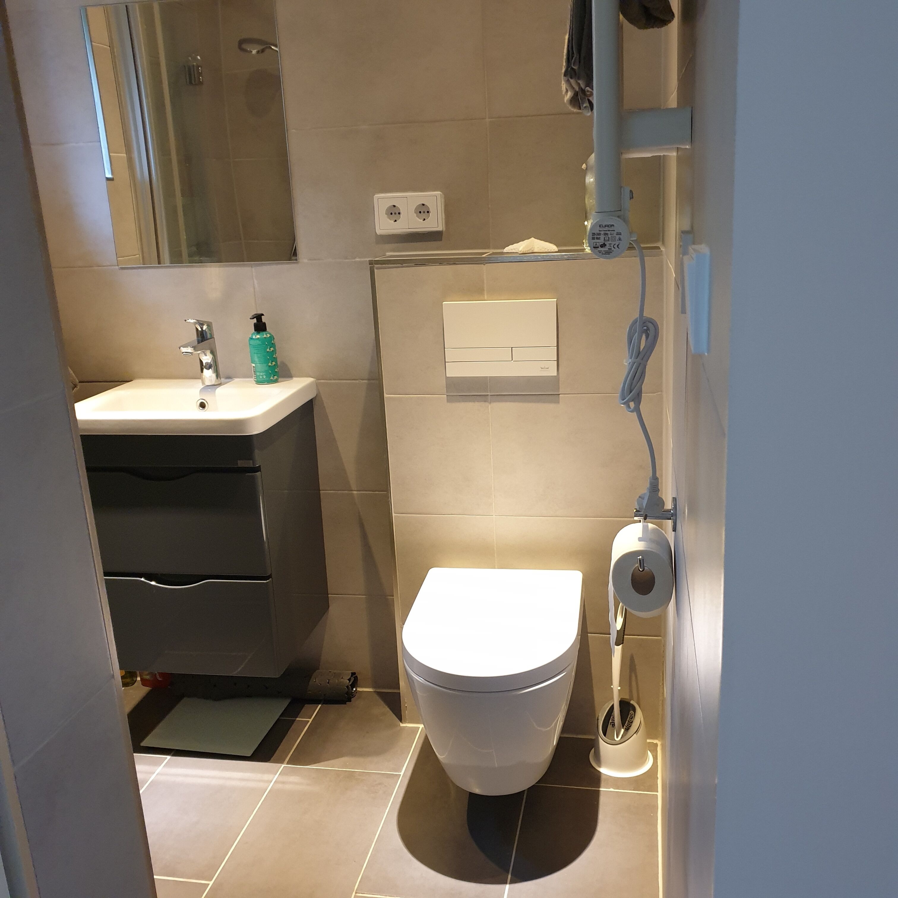 Un baño moderno con un inodoro blanco, un lavabo con grifo y un espejo. Hay una toalla colgada en la pared y un dispensador de papel higiénico.
