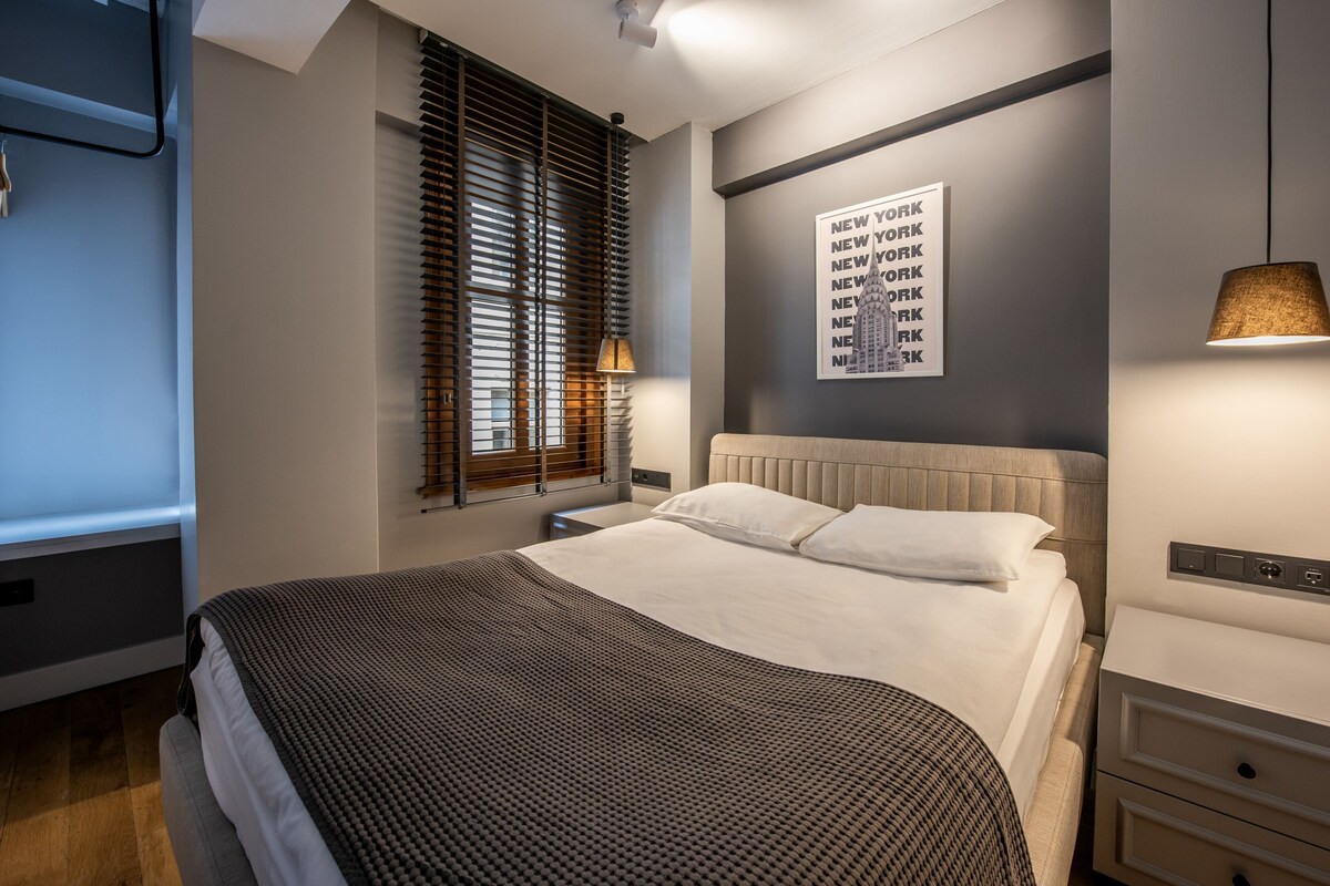 A modern hotel room with a large bed, a nightstand, a lamp, a wall-mounted artwork, and a window with blinds.