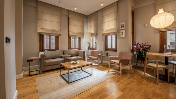 Un salón amplio con sofá, mesa de centro y sillas, suelo de madera, ventanas grandes con persianas y un comedor con mesa y sillas.