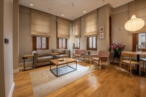 A spacious living room with a sofa, coffee table, and chairs, wooden flooring, large windows with blinds, and a dining area with a table and chairs. - Bguest Pera Suites (Istanbul)
