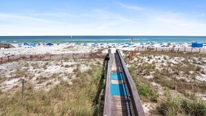 A beach with a wooden boardwalk leading to the shore, surrounded by sand dunes and beach umbrellas. - Beachfront - 2 Kings - Heated Pool - Hot tub - Low Density (Orange Beach)