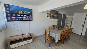 A dining area with a TV mounted on the wall, wooden chairs, a table with a striped tablecloth, and a clock on the wall. - Cosy gîte for 2 to 11 people near Gerardmer (Granges-Aumontzey)
