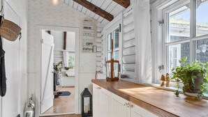 Une cuisine aux murs en bois peints en blanc, un plan de travail en bois et une fenêtre donnant sur un paysage verdoyant. On y trouve deux lanternes et une plante en pot sur le plan de travail.
