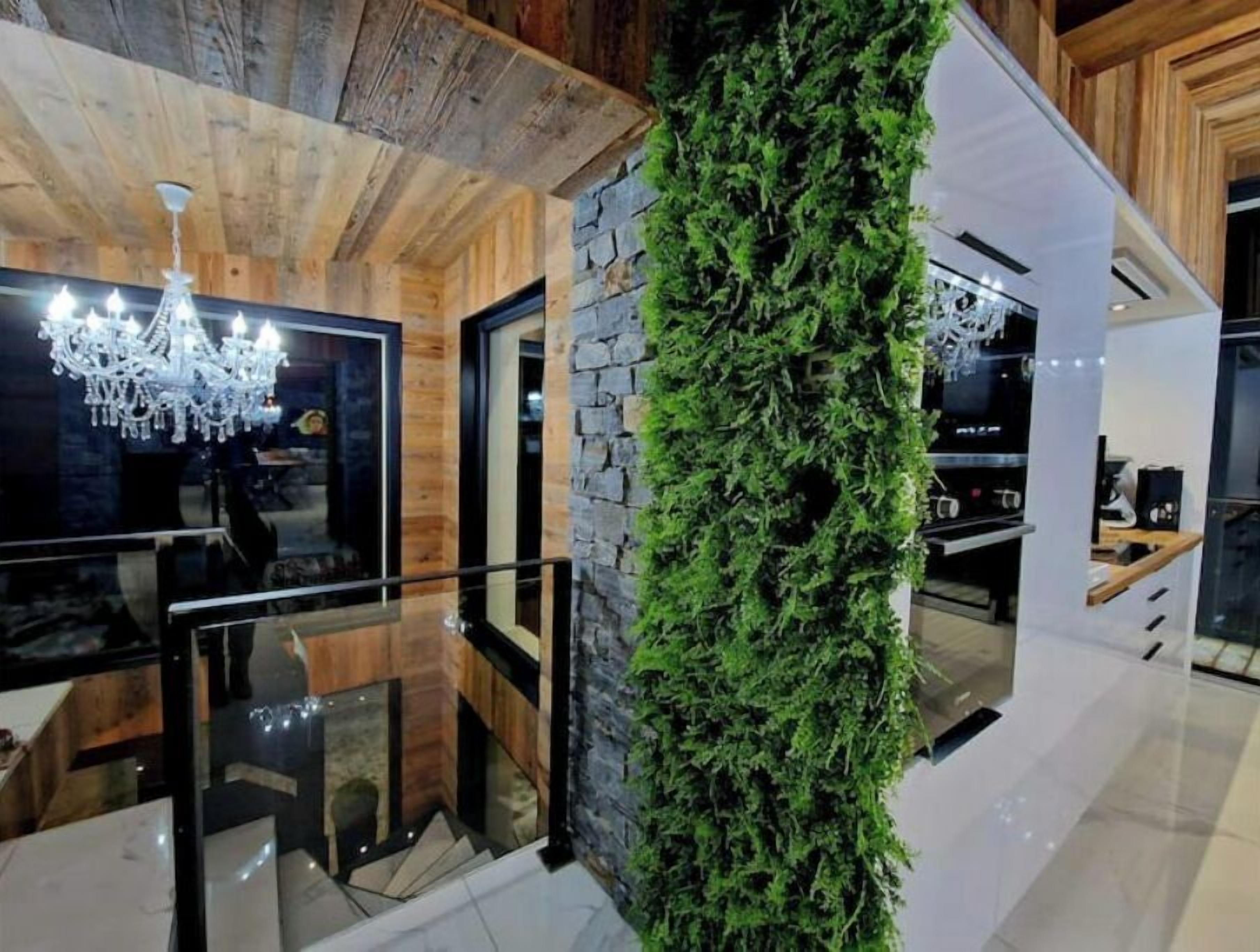 A modern interior with a wooden ceiling, a chandelier, a stone wall with greenery, and a kitchen area.