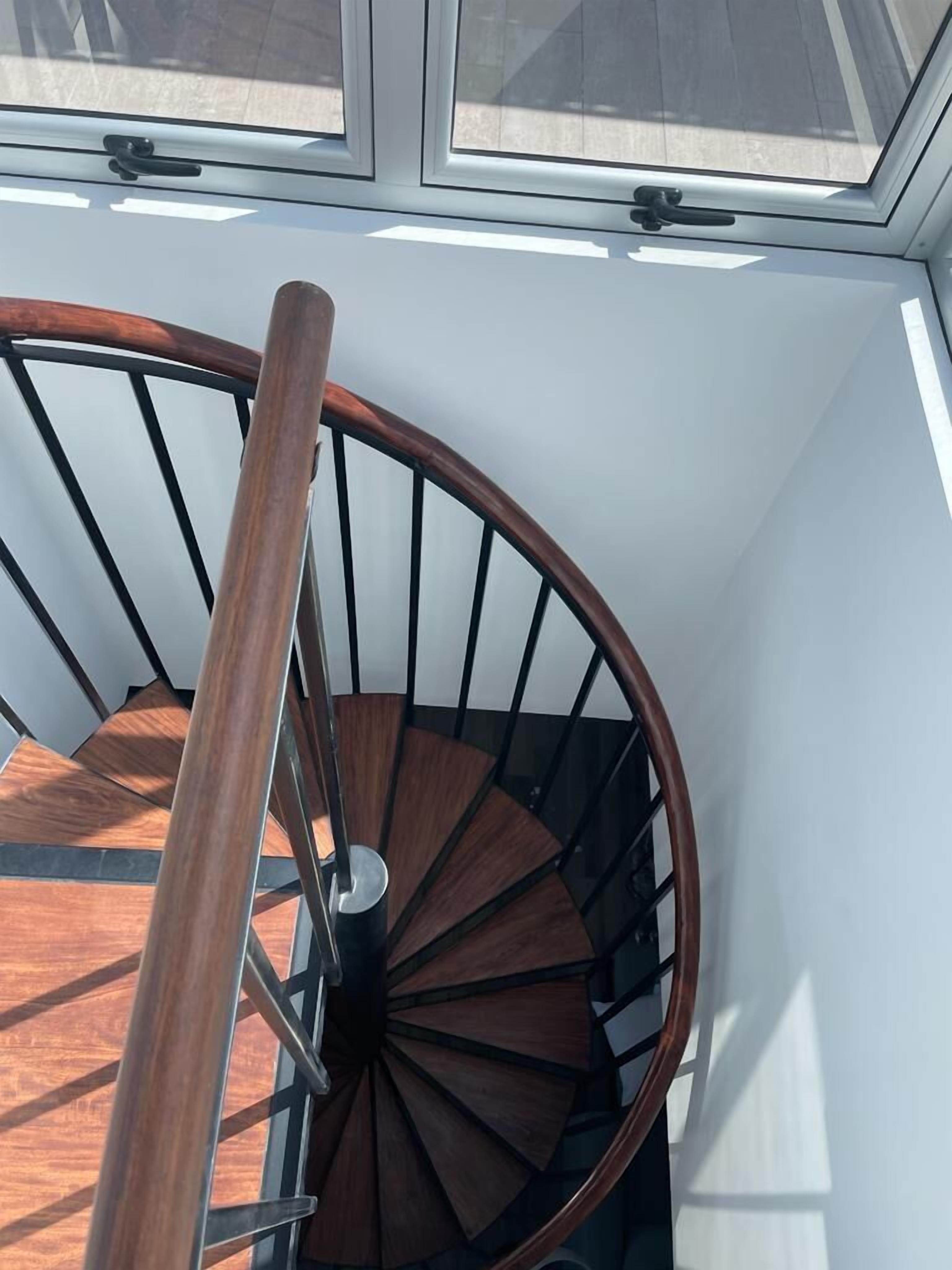 Una escalera de caracol con barandillas de madera y balaustres metálicos, que conduce a una habitación con tragaluz.