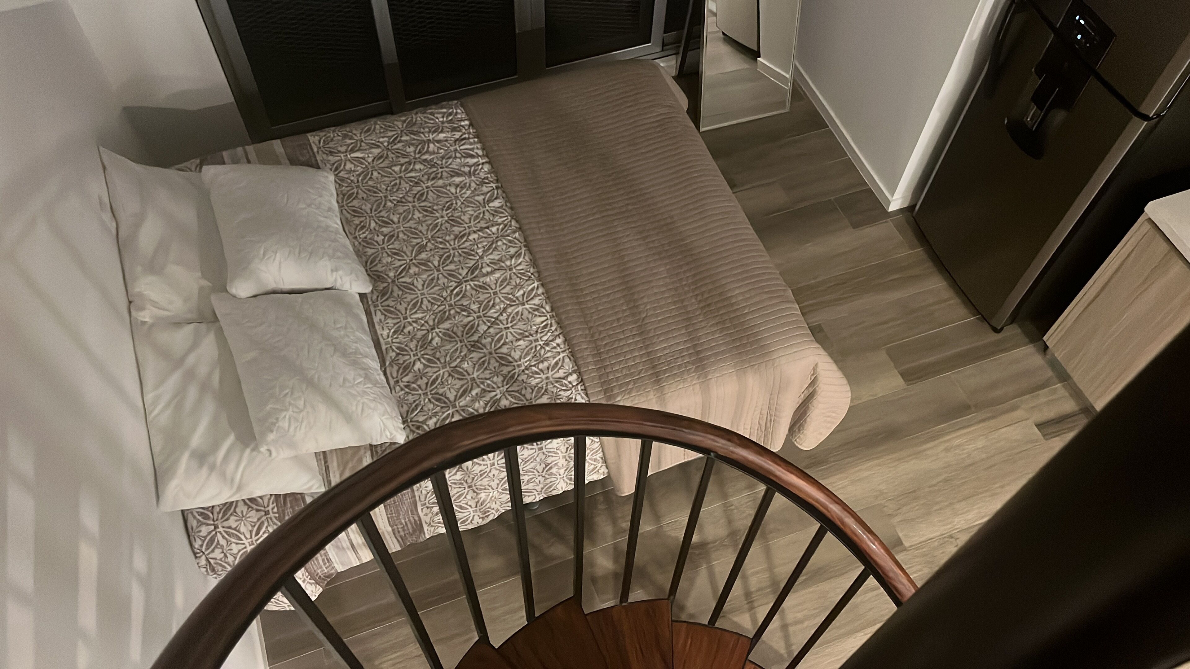 Ein Schlafzimmer mit Bett, Kissen, einer gemusterten Tagesdecke, Holzboden und einer Treppe.
