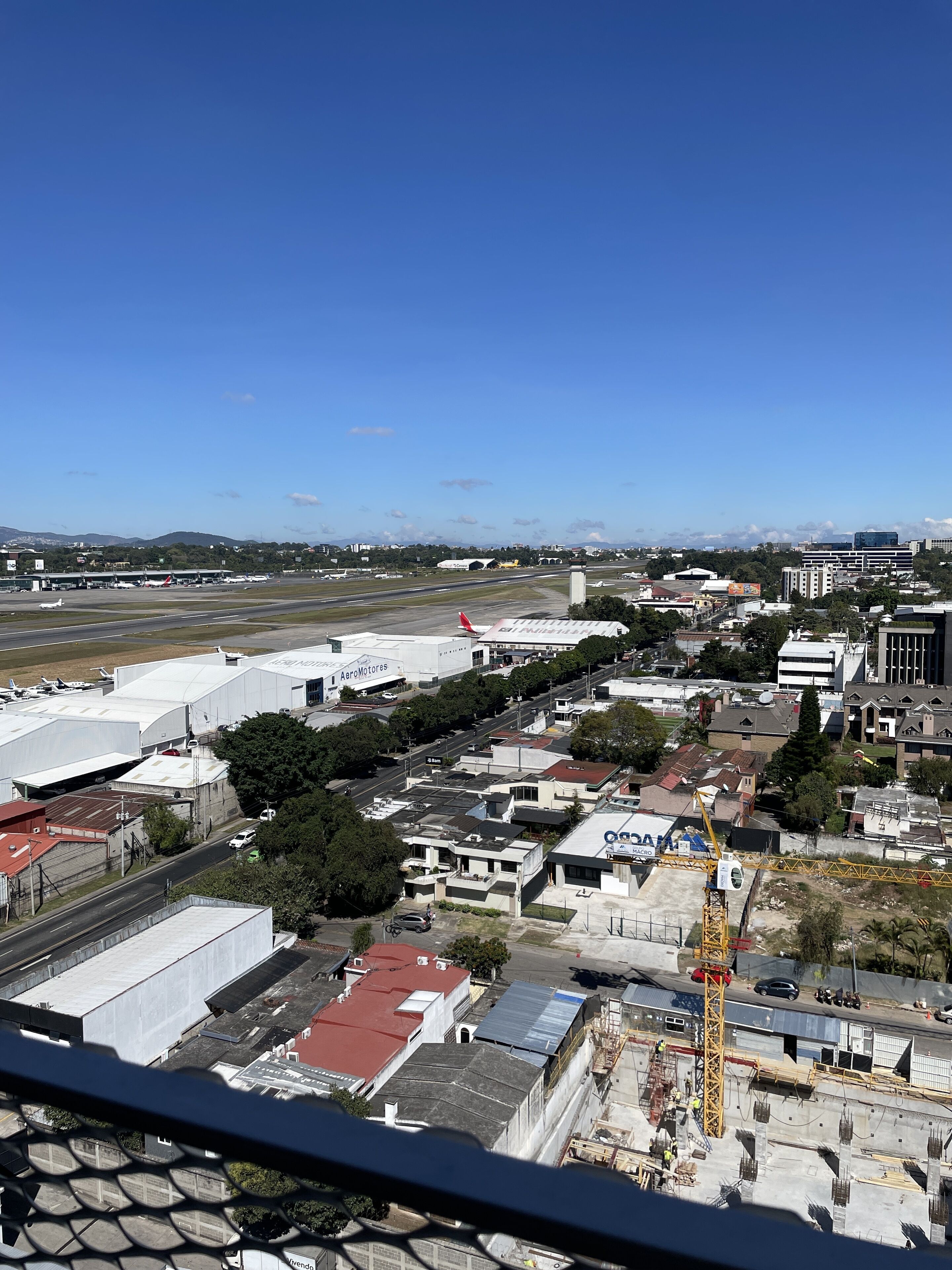 Vista aérea de un aeropuerto con varias edificaciones, incluyendo un hangar blanco grande y un edificio blanco más pequeño con la leyenda “AeroMotors”. Se observan varios vehículos en el suelo, y una grúa de construcción es visible en primer plano.