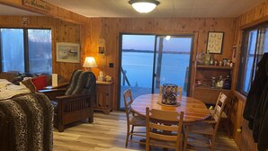 L’intérieur d’une cabane avec un plancher en bois, une table à manger ronde, un canapé, une lampe et une vue sur un lac, visible à travers de grandes fenêtres.