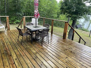 Eine Holzterrasse mit Tisch und Stühlen, umgeben von einem Hollerdgeschoss und Blick auf einen See.