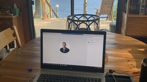 Un ordinateur portable est posé sur une table en bois, et une porte vitrée offre une vue sur un lac et des arbres.