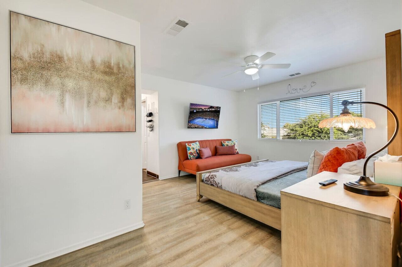 A bedroom with a bed, a sofa, a ceiling fan, a TV, and a window with blinds.