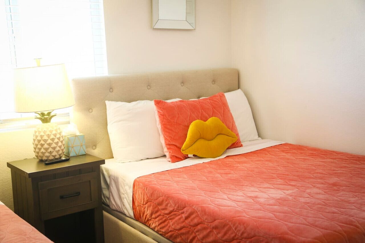 A neatly made bed with a red quilt, a beige headboard, and two pillows, one white and one orange.