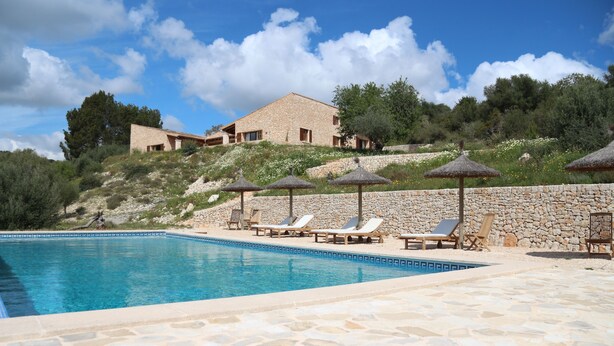 Une piscine entourée de chaises longues et de parasols, avec des murs en pierre et un bâtiment en pierre en arrière-plan.