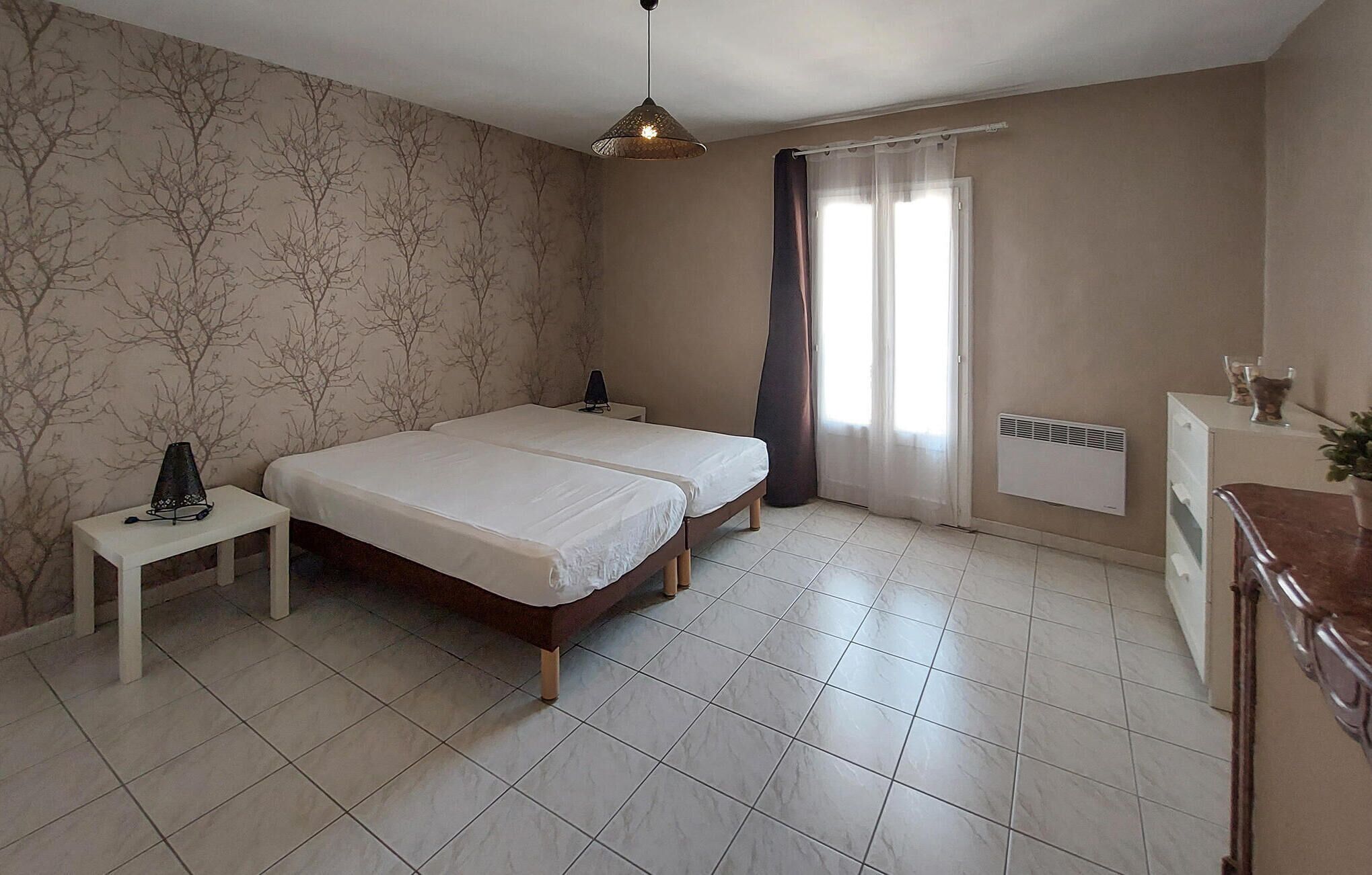 Une chambre avec deux lits, une table de chevet, une lampe, un radiateur et une fenêtre avec des rideaux.