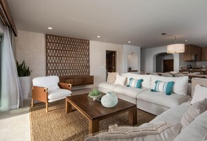 Un salon moderne avec un canapé, un fauteuil et une table basse. On aperçoit un coin cuisine au fond.