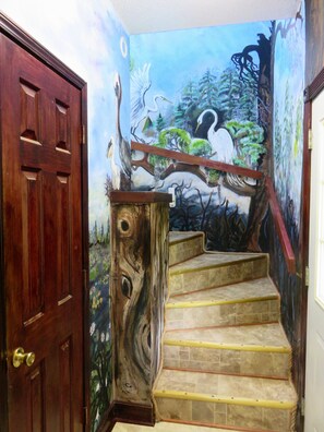 Une porte en bois avec une poignée en laiton, un escalier doté d’une rampe et une fresque représentant un oiseau et des arbres.