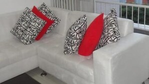 A white sofa with patterned cushions and a red accent cushion, positioned in a room with a large window and a view of buildings outside. - Emdoralahouse (Cali)