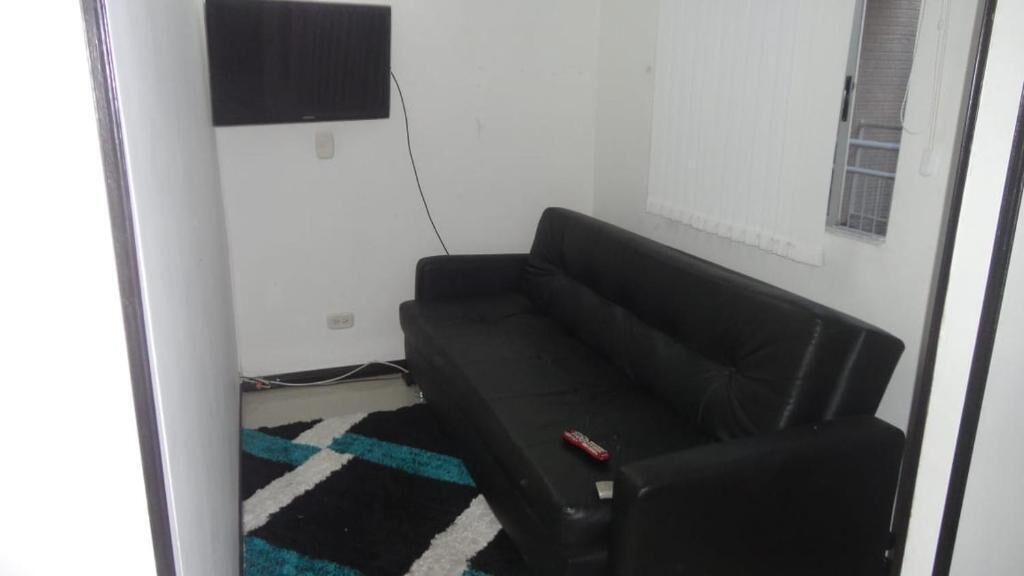 Een kleine, eenvoudig ingerichte kamer met een zwarte bank, een kamerkleed met een patroon en een raam met jaloezieën.