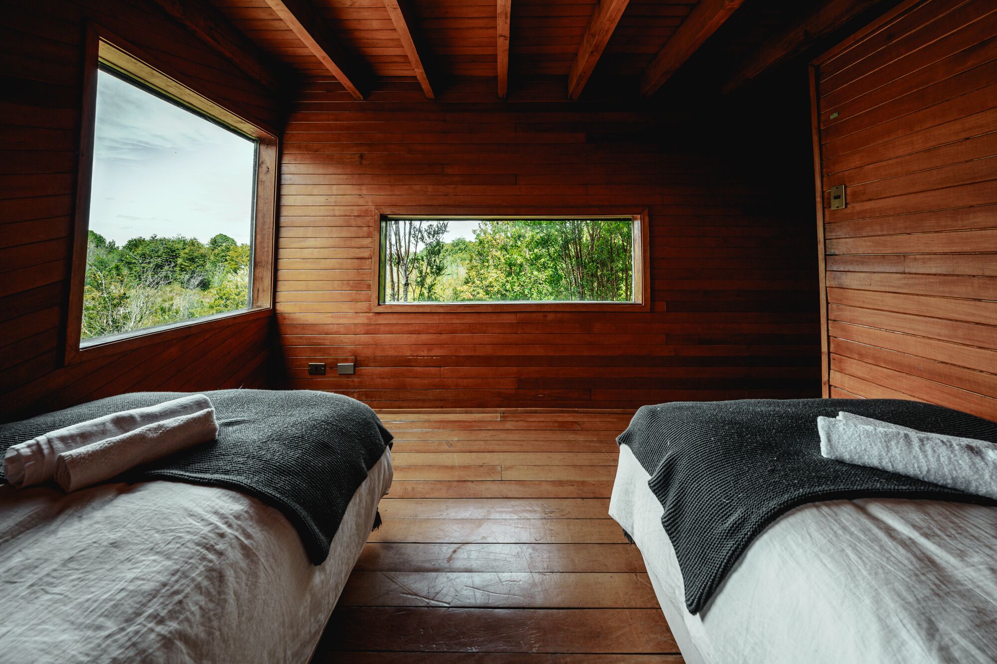 A cabin with two beds, wooden walls, a window with a forest view, and a wooden floor.