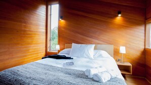 A wooden-paneled bedroom with a bed, white bedding, a nightstand with a lamp, and a window providing natural light. - Hotel Parque Tepuhueico (Chonchi)