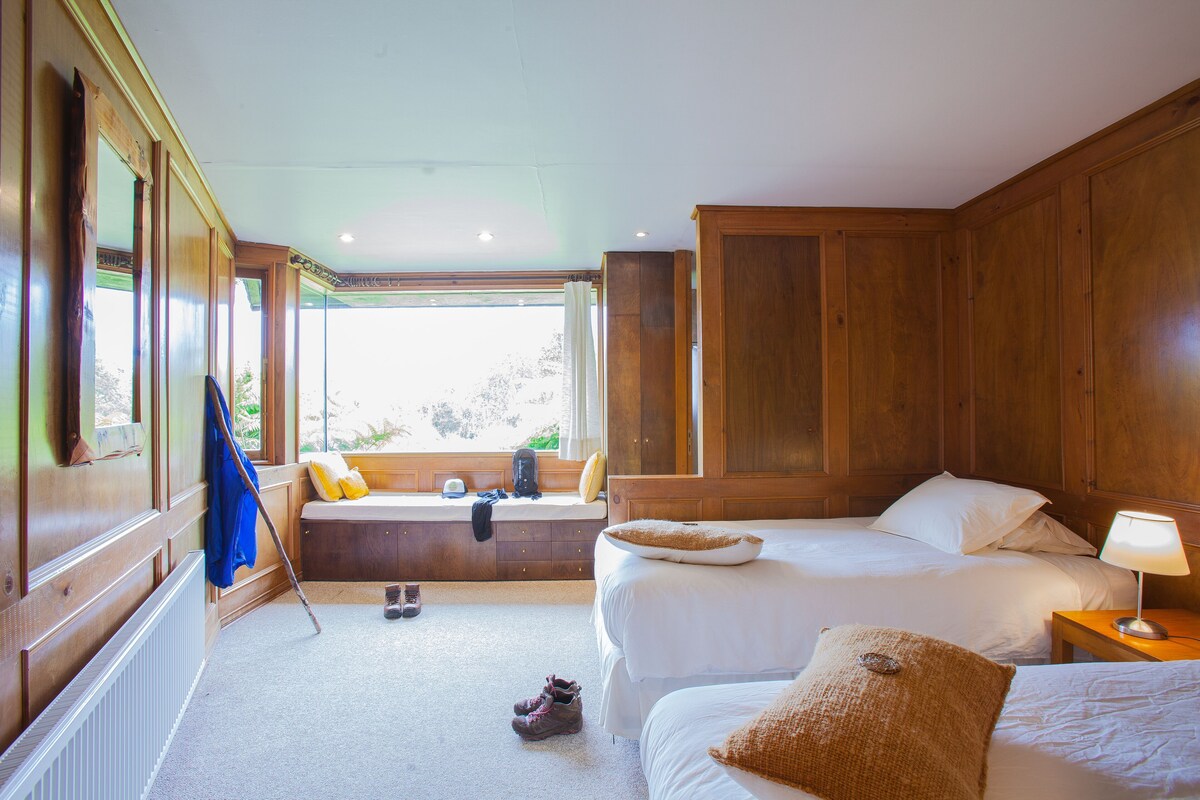 A room with two beds, a window seat, wooden walls, and a radiator.