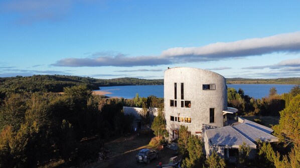 Un bâtiment moderne en pierre, doté d’une tour circulaire, entouré d’une forêt dense et d’un lac.