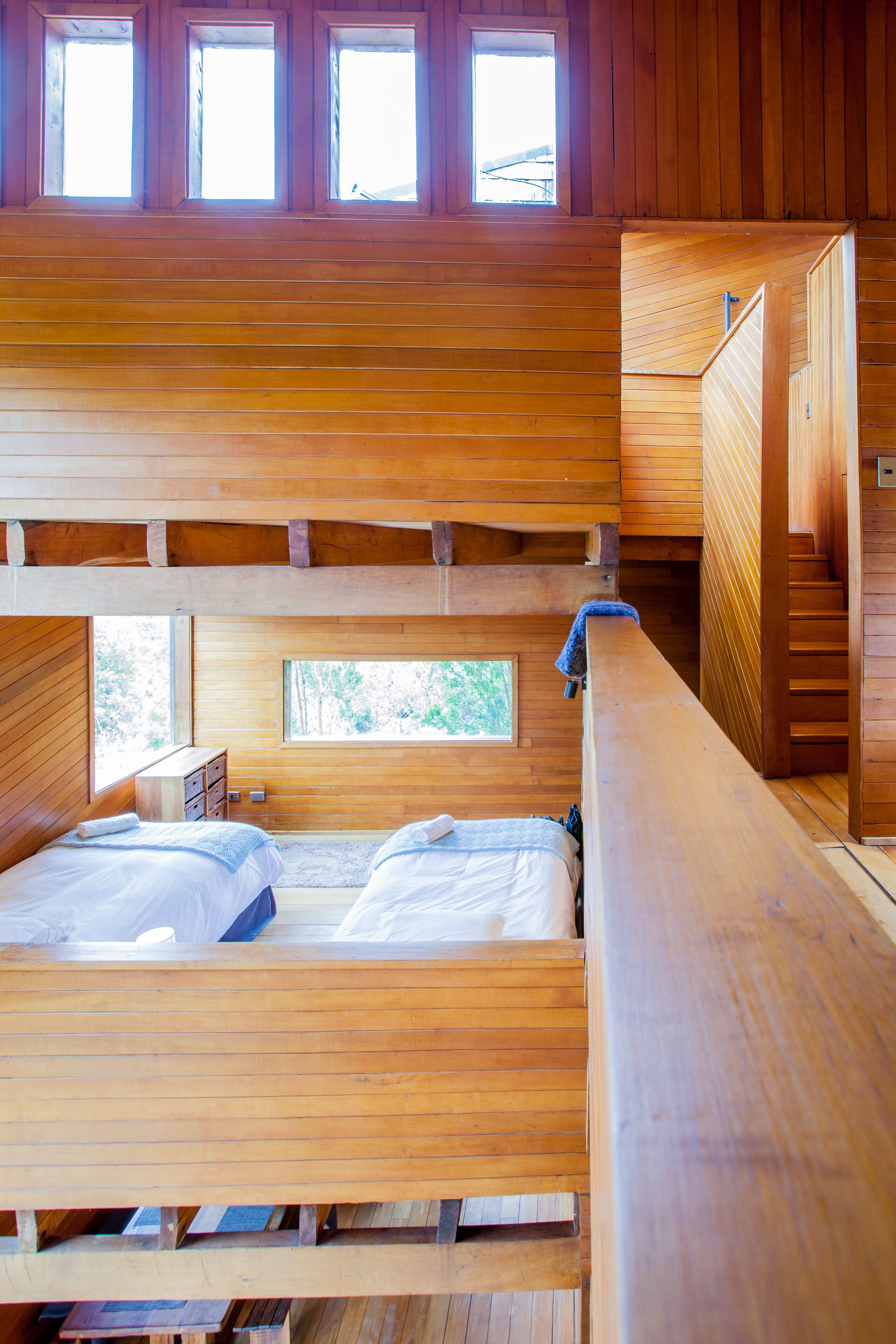 A cabin with two wooden beds, a small wooden desk, and a window with a view of trees.