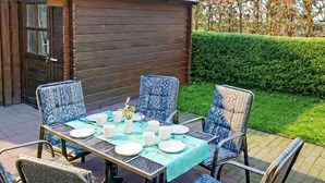 Un área de comedor al aire libre con una mesa dispuesta para cuatro personas, sillas con diseños llamativos, un edificio de madera al fondo y un seto bien cuidado.