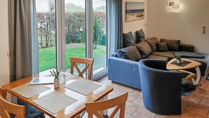 Una sala de estar moderna con un sofá, una mesa de comedor con sillas y vistas a un jardín a través de amplios ventanales.