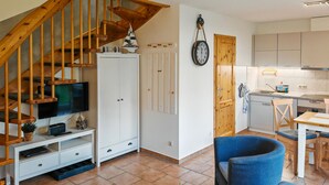 Una sala moderna con una escalera de madera, un televisor sobre un mueble blanco, un comedor con una silla azul y una cocina con armarios blancos.