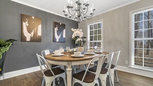 A dining area with a round table set for six, surrounded by white chairs with dark cushions. There is a chandelier above the table, and artwork is displayed on the walls.