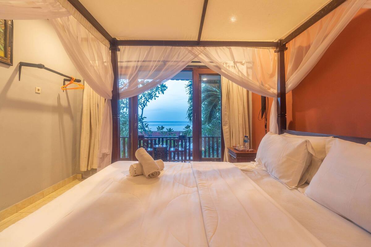 Un dormitorio con una cama grande, un balcón con vistas al mar y una ventana con cortinas translúcidas.