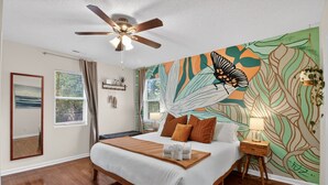 Una camera da letto con un letto grande, una ventola a soffitto, una finestra con tende, un affresco e un comodino con una lampada.