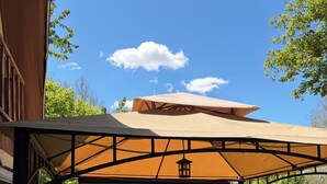 Una terraza de madera con un porche cubierto, barandilla metálica y vistas a un jardín con árboles y arbustos.