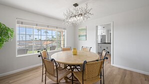 A dining area with a round table, six wicker chairs, a chandelier, and large windows with a view of palm trees.