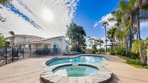 A swimming pool with a stone edge, a covered patio area with lounge chairs, and a house with a tiled roof.