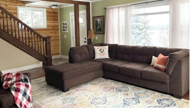 Un salon avec un canapé marron, un pouf assorti, un tapis à motifs, un ventilateur de plafond, un escalier et une grande fenêtre avec des rideaux.