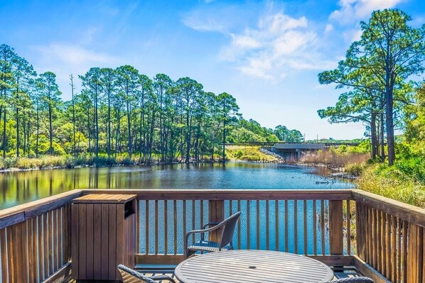 A wooden deck with a table and chairs overlooking a calm river, surrounded by dense greenery and a clear sky.