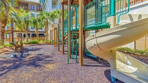 A playground with slides and climbing structures, surrounded by palm trees and a building with balconies. - Redfish Village - Beach Please - 2BR/2BA - Gulf Views - Seasonal Beach Tram (Santa Rosa Beach)