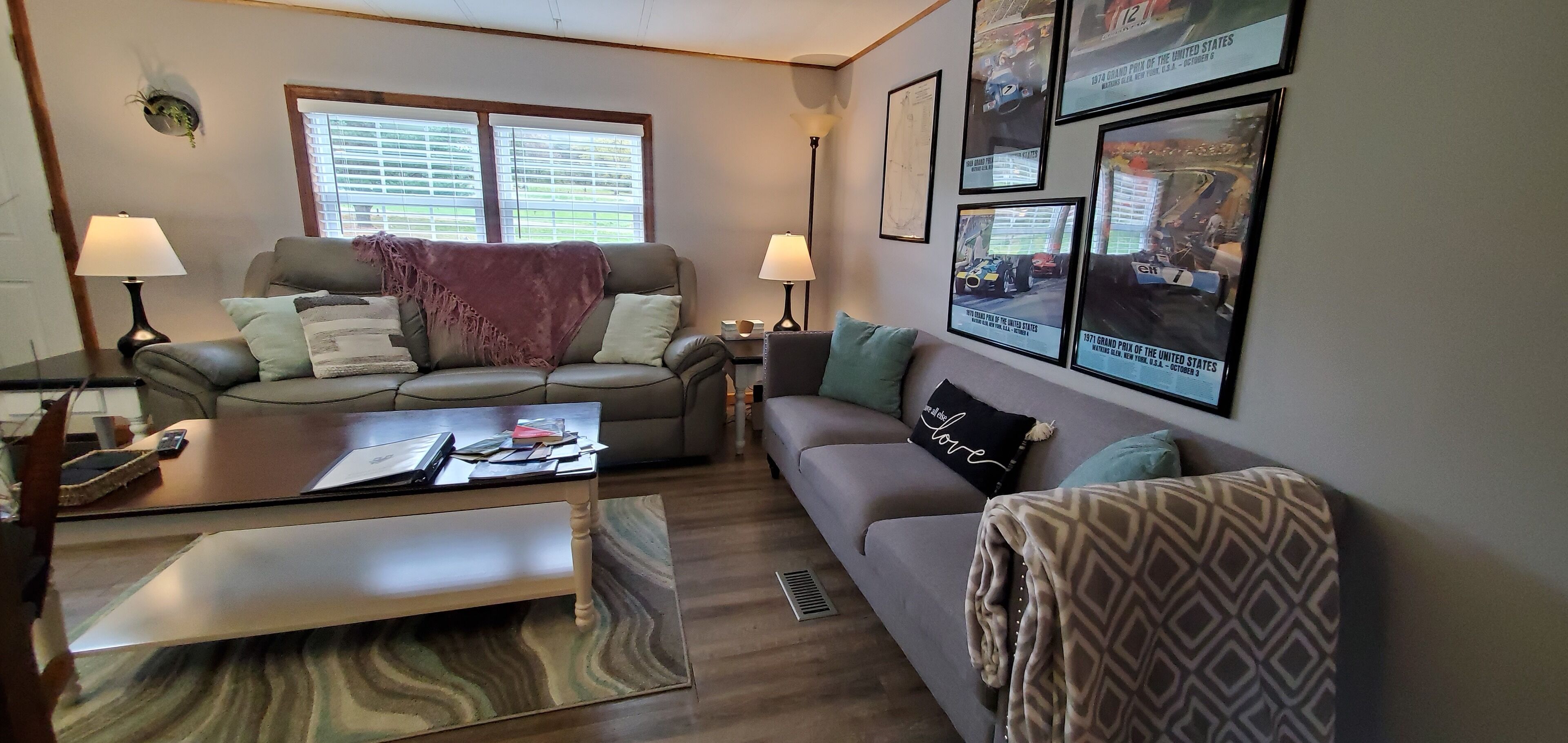 A living room with two sofas, a coffee table, and a TV. There are framed pictures on the wall and a window with blinds.