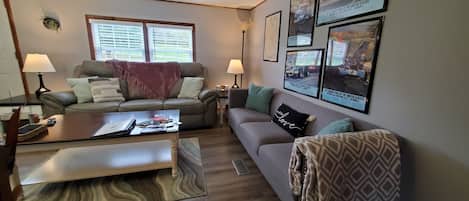 A living room with two sofas, a coffee table, and a TV. There are framed pictures on the wall and a window with blinds.