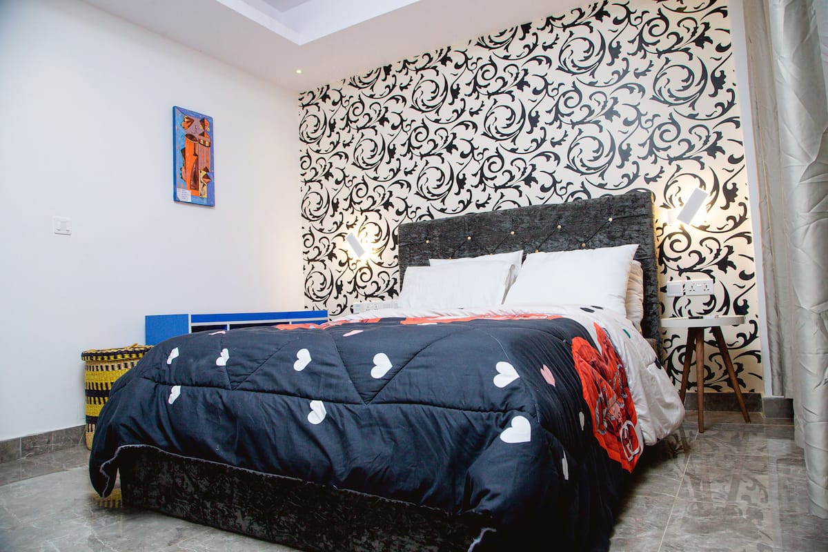 Un dormitorio con cama, papel tapiz estampado, una mesita pequeña y una lámpara de pared.