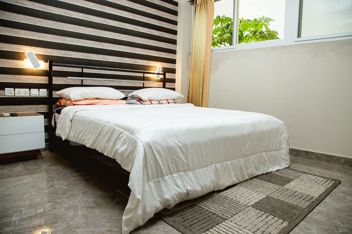 Una cama bien hecha con ropa de cama blanca, una mesita de noche con una lámpara y una ventana con cortinas.