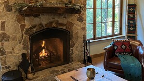 Una chimenea de piedra con un televisor encima, un sofá de cuero con una manta, y una ventana con vistas a árboles.