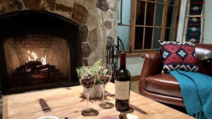 Una sala de estar acogedora con chimenea, un sofá de cuero y una mesa de madera con una botella de vino, copas y una tabla de embutidos.