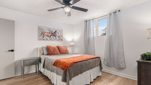 Ein Schlafzimmer mit Bett, Deckenventilator, Nachttisch, Lampe und Fenster mit Vorhängen.