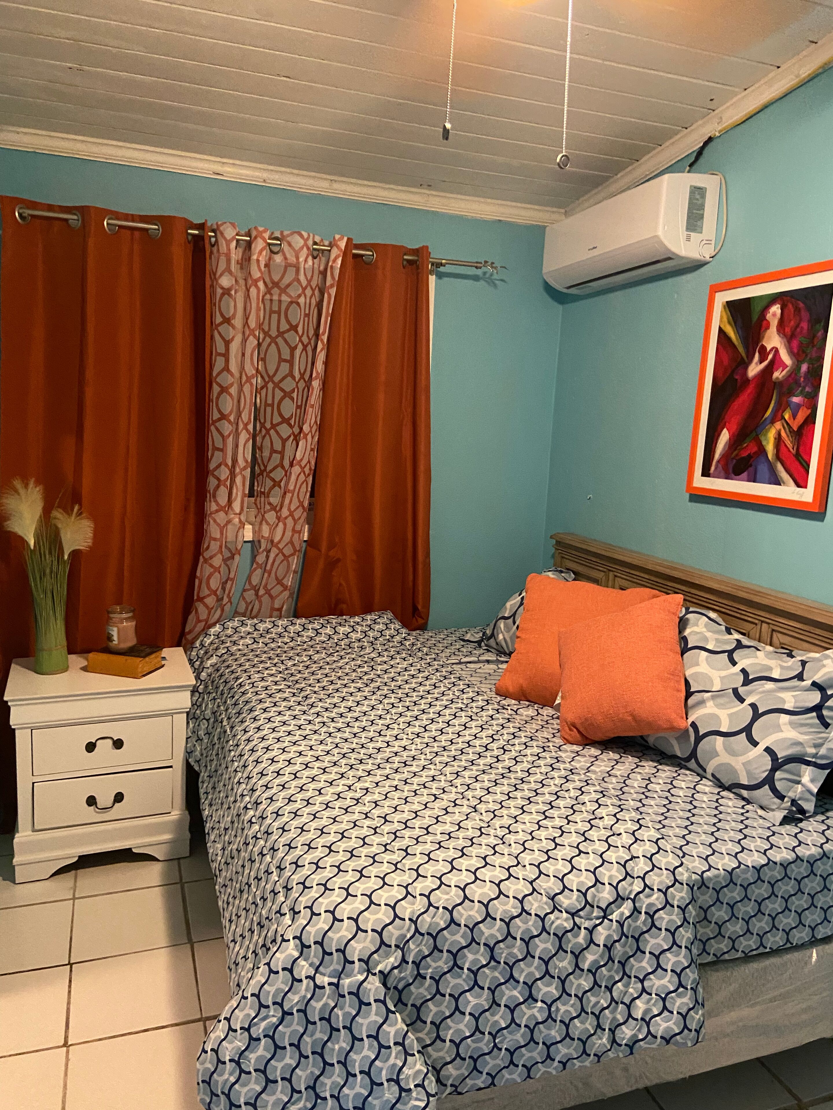 A bedroom with a bed, a nightstand, a wall-mounted air conditioner, and a painting.
