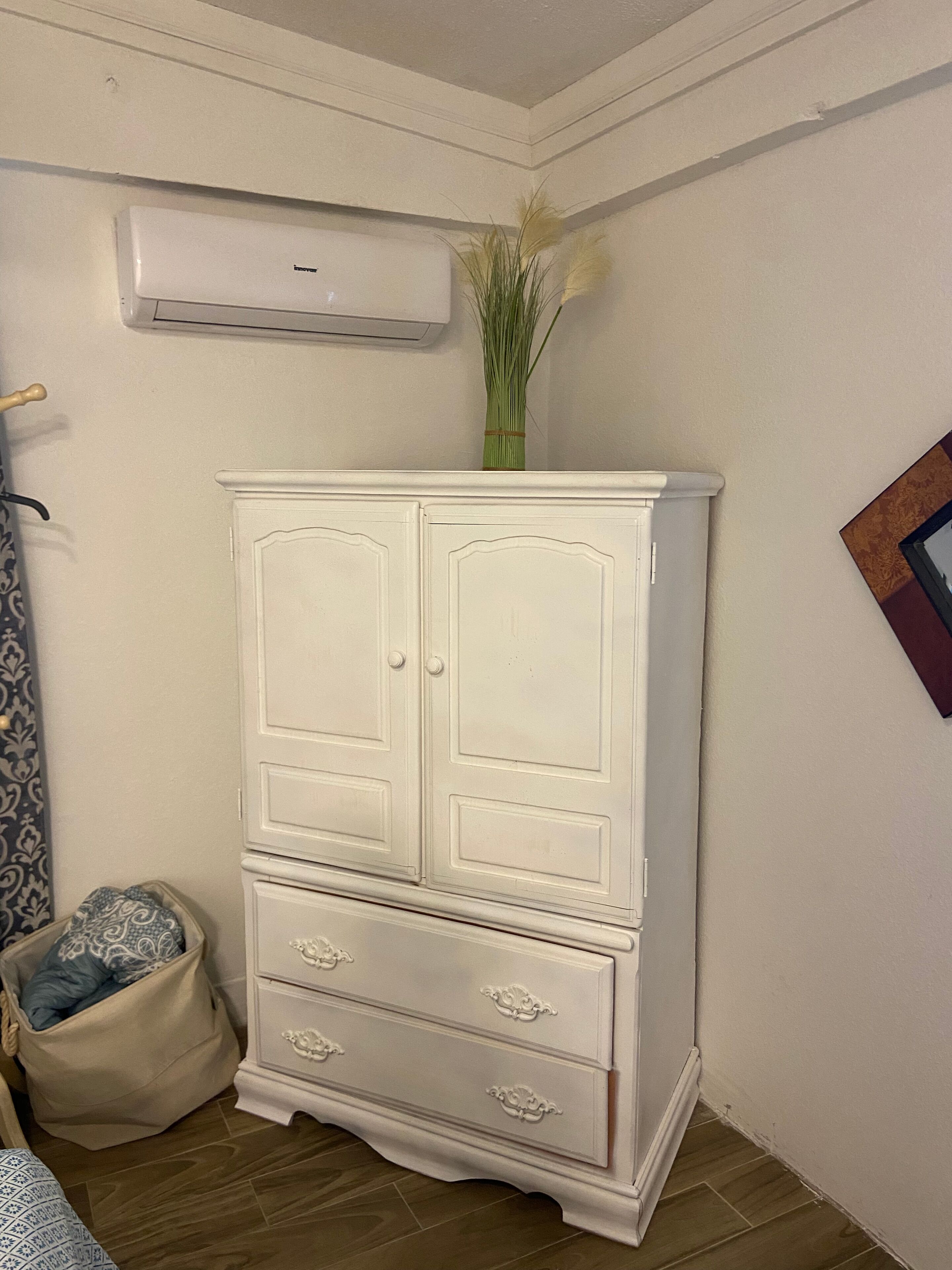 A white wardrobe with two doors and two drawers, a small plant on top, and a wall-mounted air conditioner.