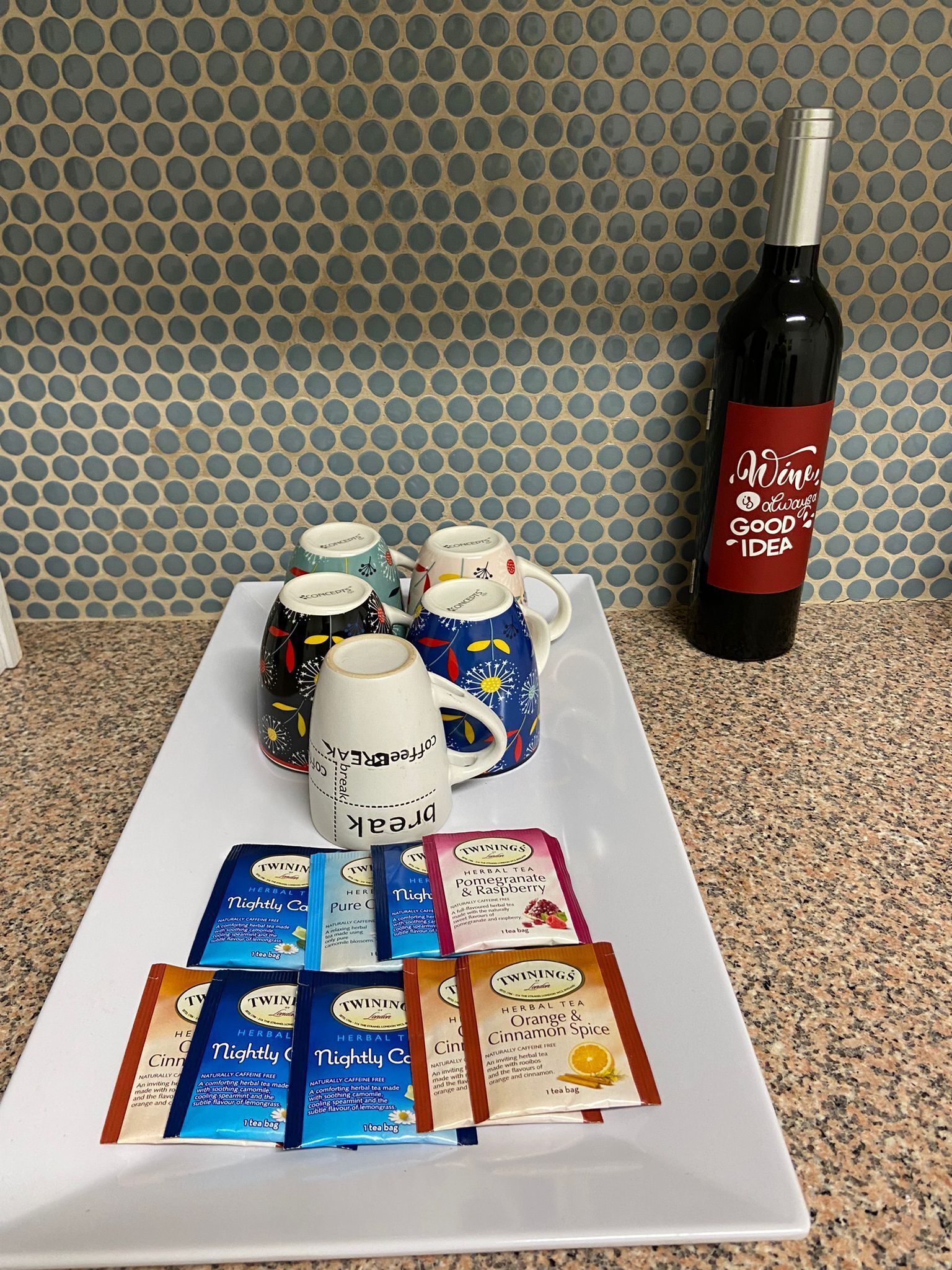 A tray with Twinings herbal tea bags, a bottle of wine, and decorative mugs on a granite countertop.